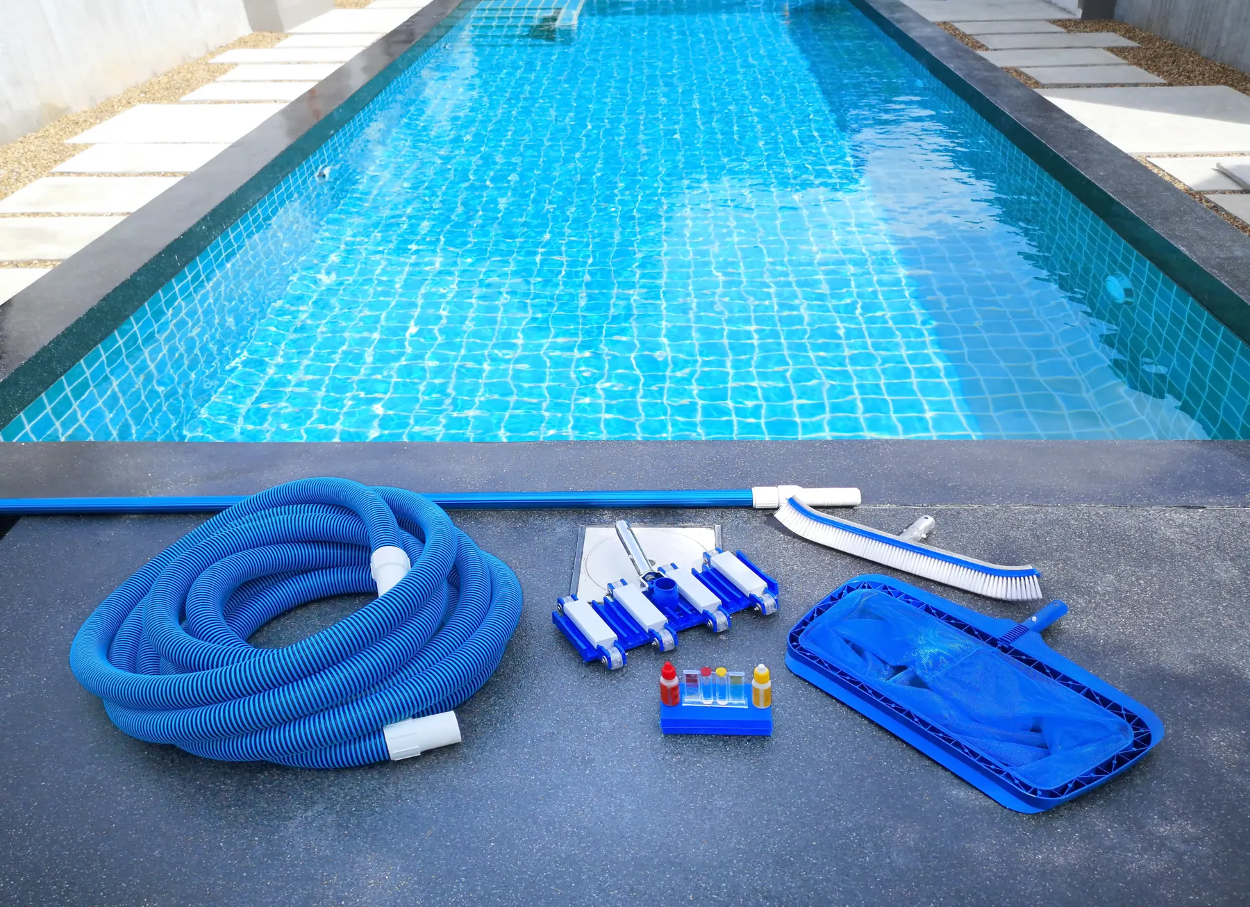 Pool cleaning equipment -- a vacuum hose, skimmer net, brush, and water-testing kit -- laid out poolside
